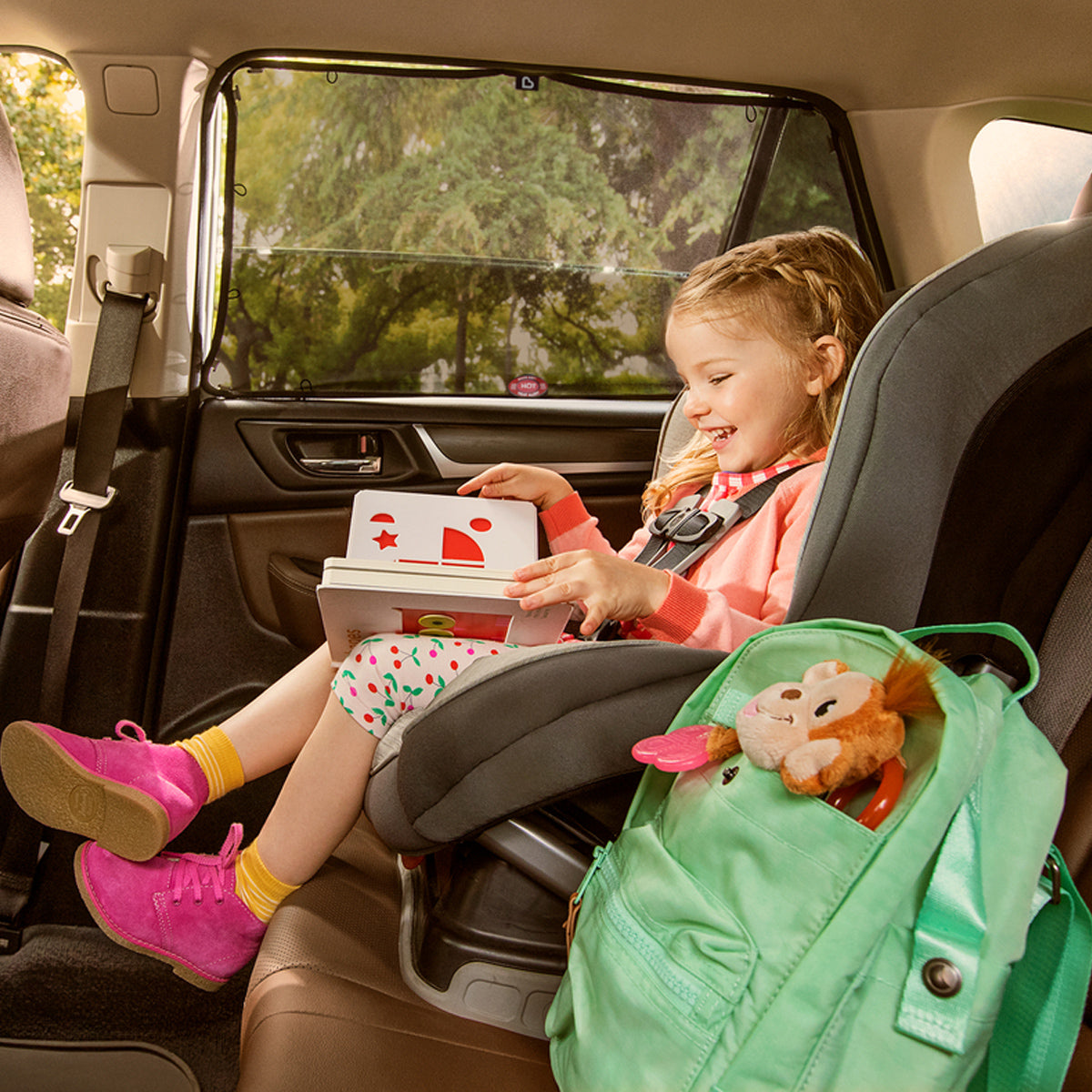Young girl in a car seat reading a book with the Munchkin sun shade fitted to the rear window beside her