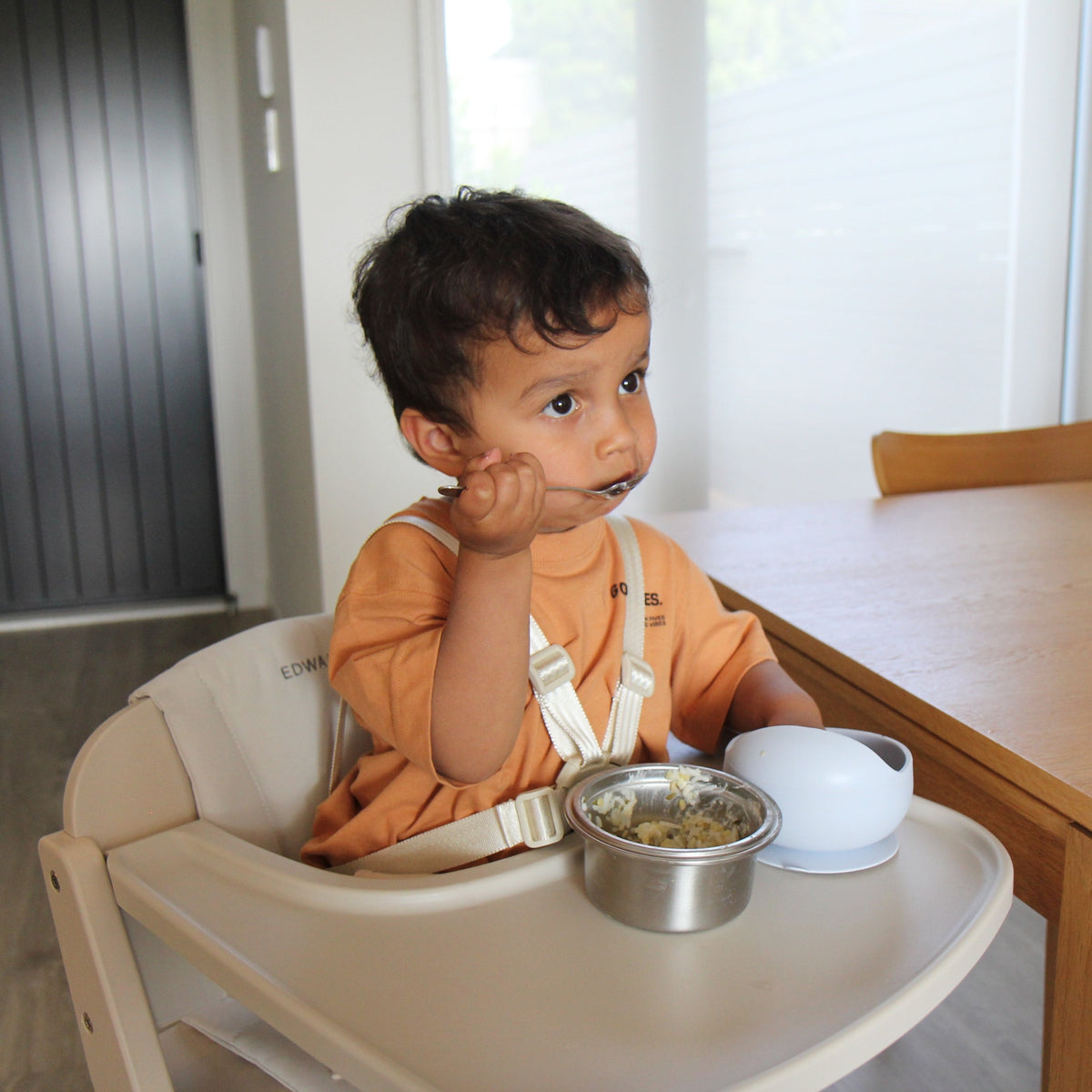 Toddler eating from a bowl in the Cariboo Frankie Highchair pulled up to the family dining table (shown in Oat)