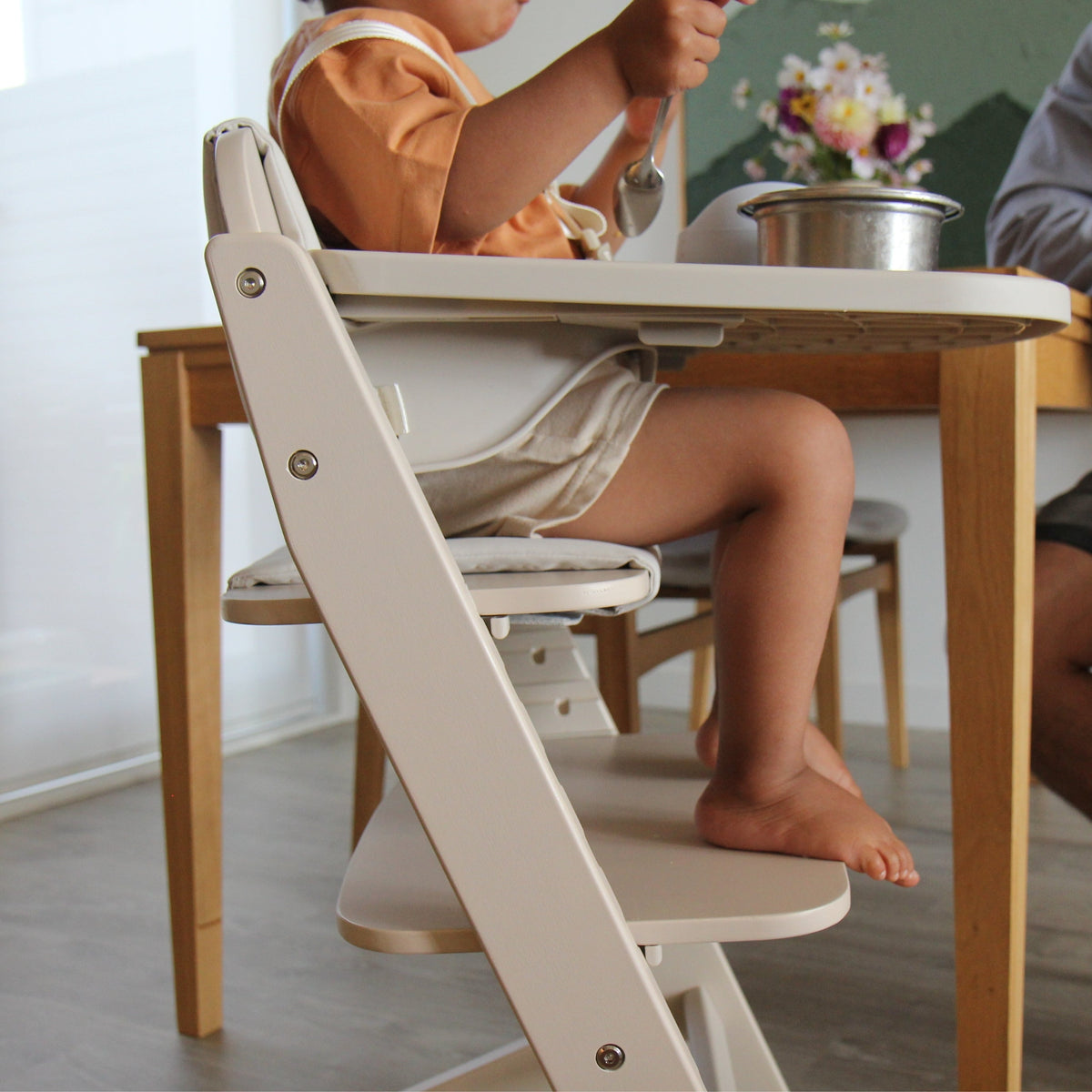 Side view of the Cariboo Frankie Highchair with toddler seated at the family table, tray removed (shown in Oat)