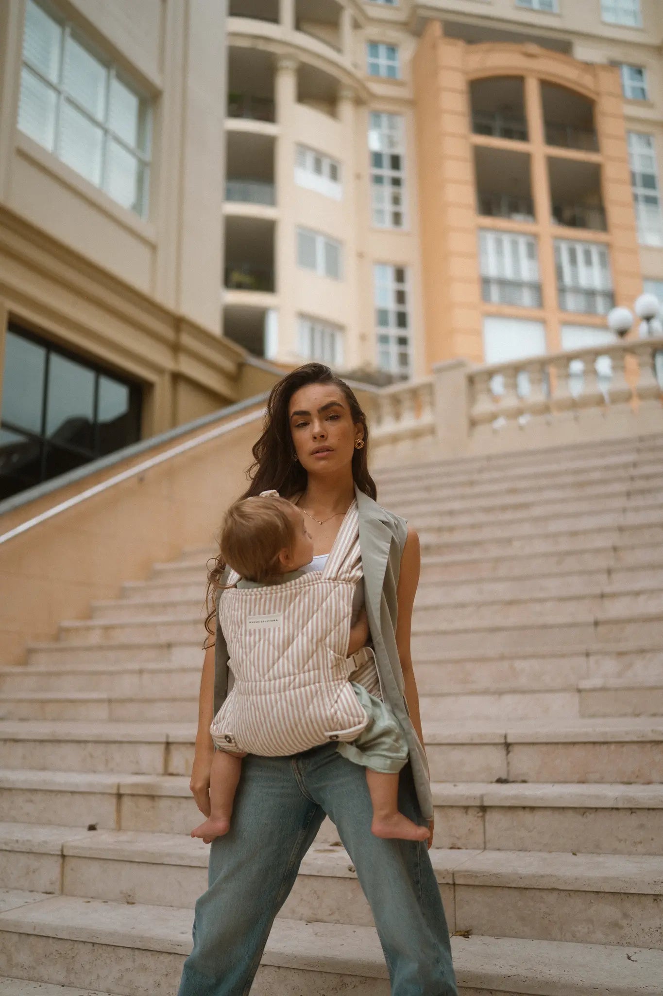 Mum carries her toddler in the Burleigh stripe Daily Carrier on city stone steps