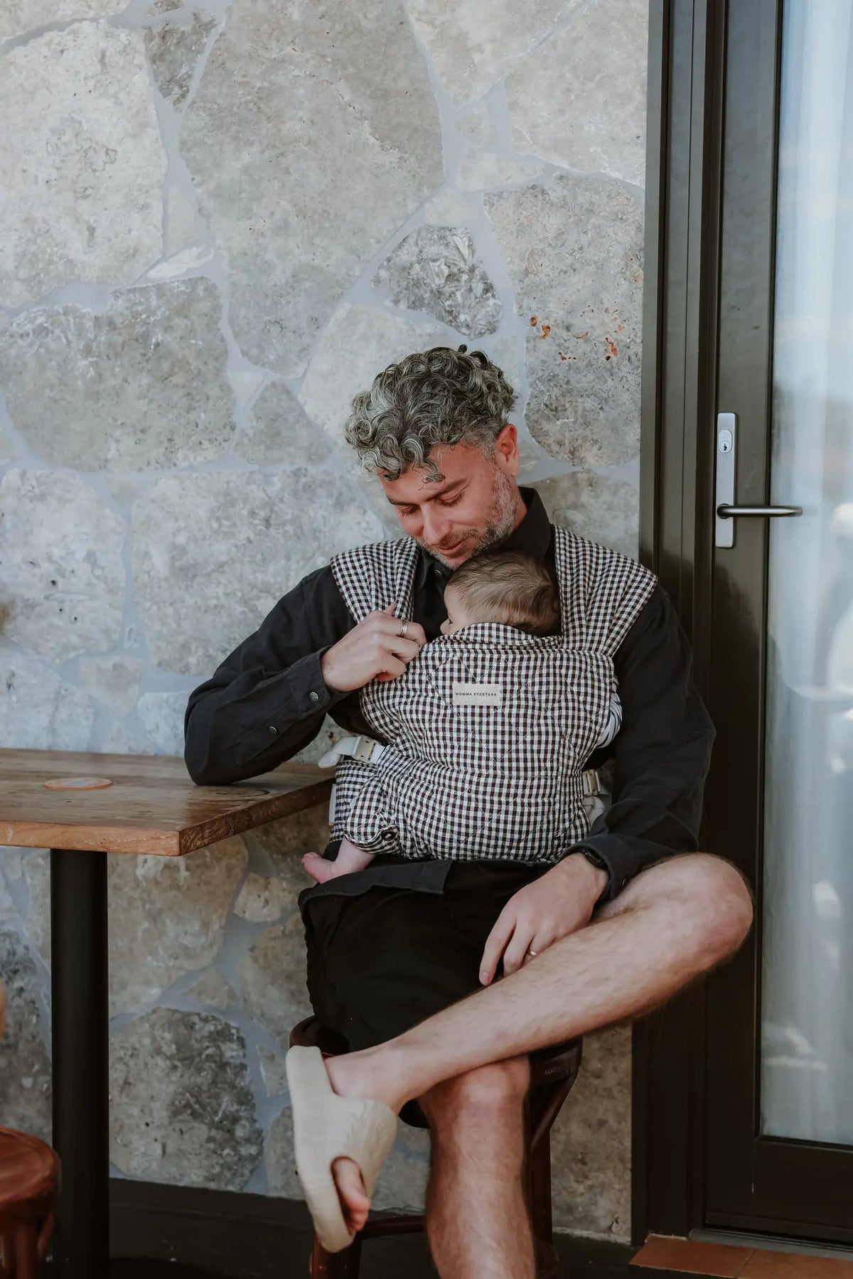 Dad settles his sleeping baby into the Elston gingham Daily Carrier at a cafe table