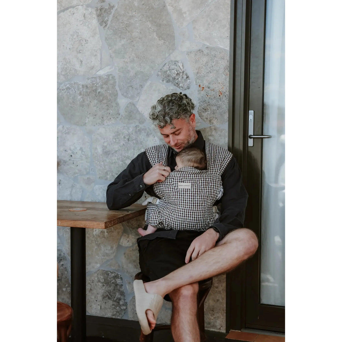 Dad settles his sleeping baby into the Elston gingham Daily Carrier at a cafe table