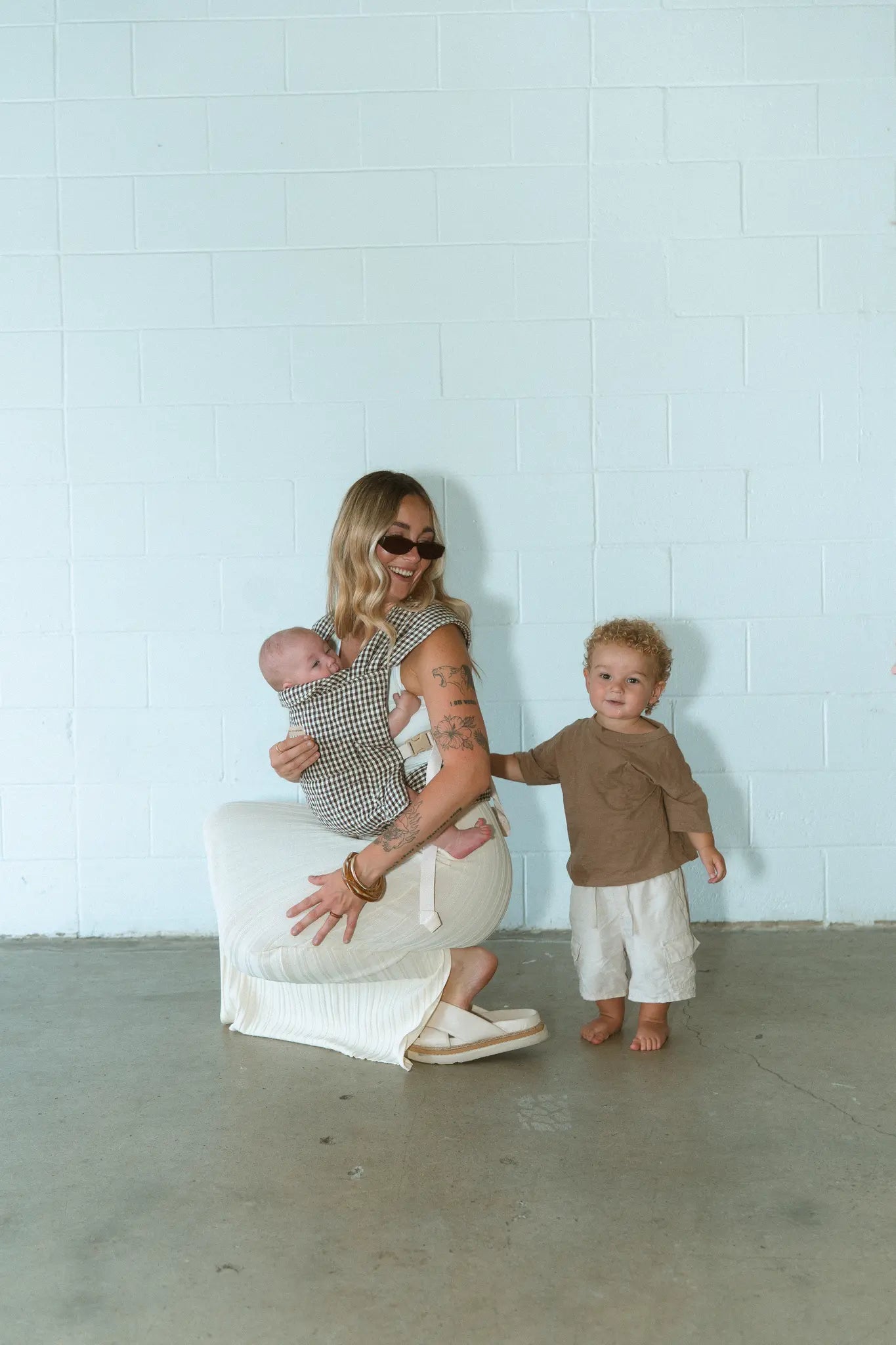 Mum crouches with her newborn in the Elston gingham Daily Carrier while her toddler stands beside her