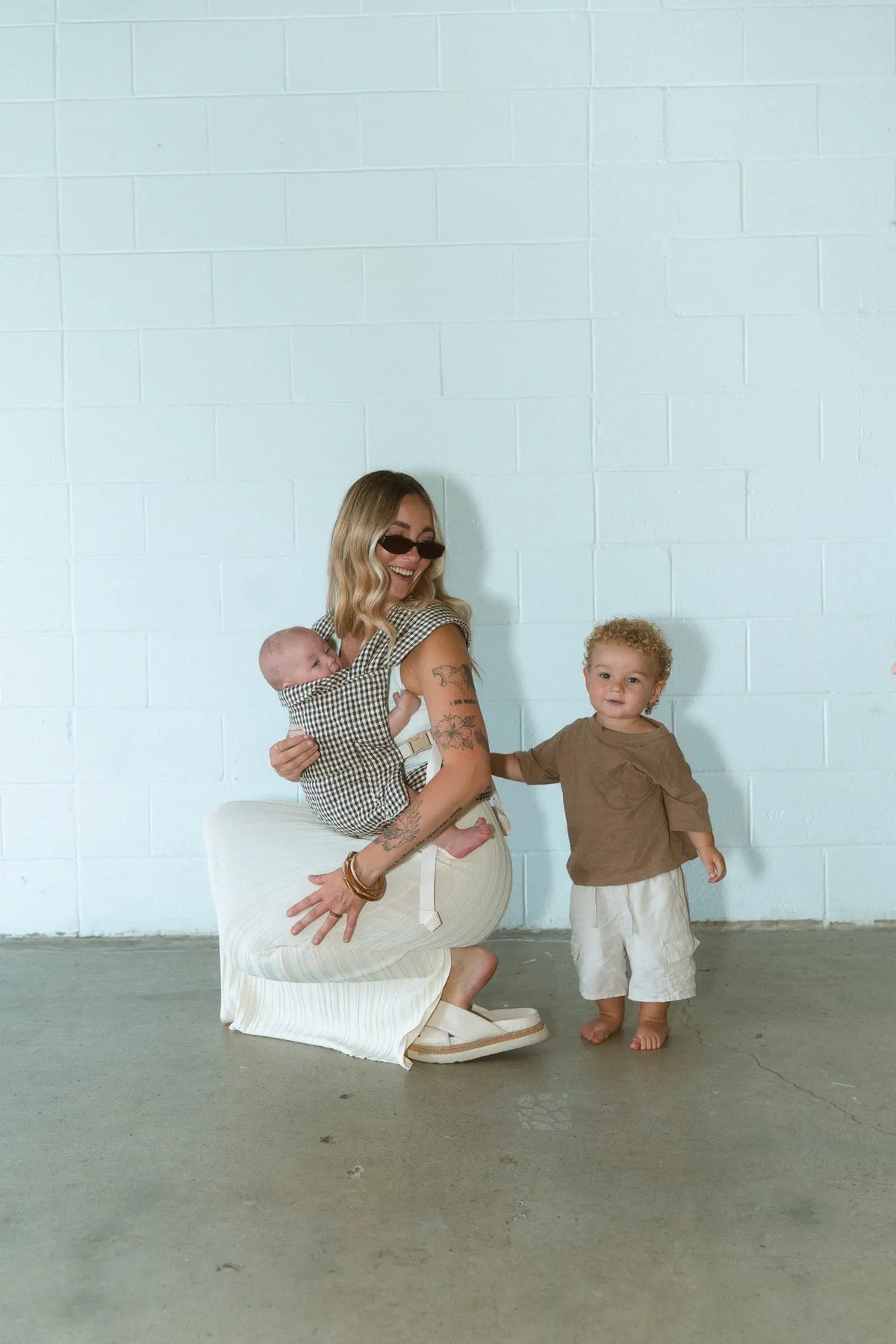 Mum crouches with her newborn in the Elston gingham Daily Carrier while her toddler stands beside her