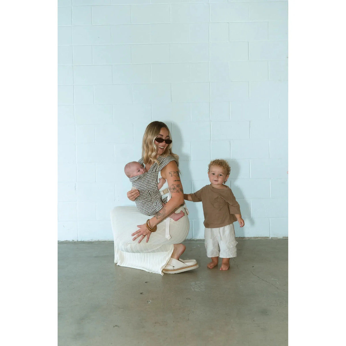 Mum crouches with her newborn in the Elston gingham Daily Carrier while her toddler stands beside her