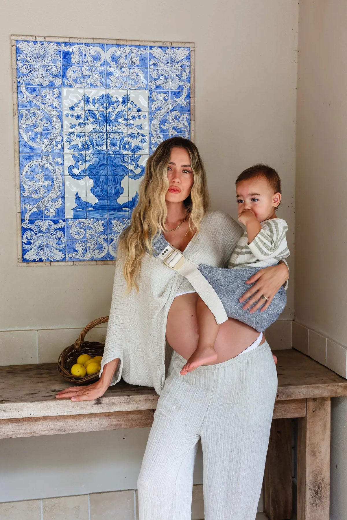 Mum leans on a bench by blue tiles, toddler held in the Lane linen Hipsy Carrier